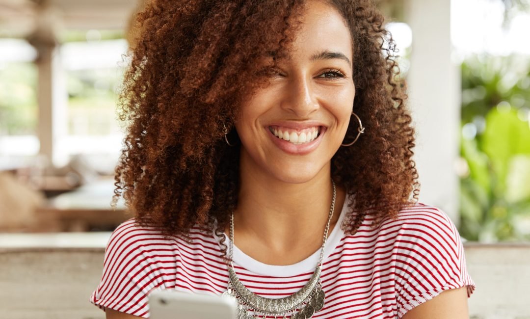 Happy smiling African American female with curly hairstyle sends feedback to interesting video on internet website, uses modern cell phone for chatting with friends duing free time in restaurant Happy smiling African American female with curly hairstyle sends feedback to interesting video on internet website, uses modern cell phone for chatting with friends duing free time in restaurant